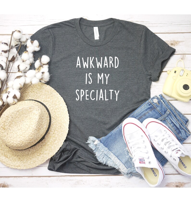 May include: A dark gray t-shirt with the text "AWKWARD IS MY SPECIALTY" in white lettering. The shirt is laid out on a white wooden surface with a straw hat, blue denim shorts, white sneakers, and a yellow camera.