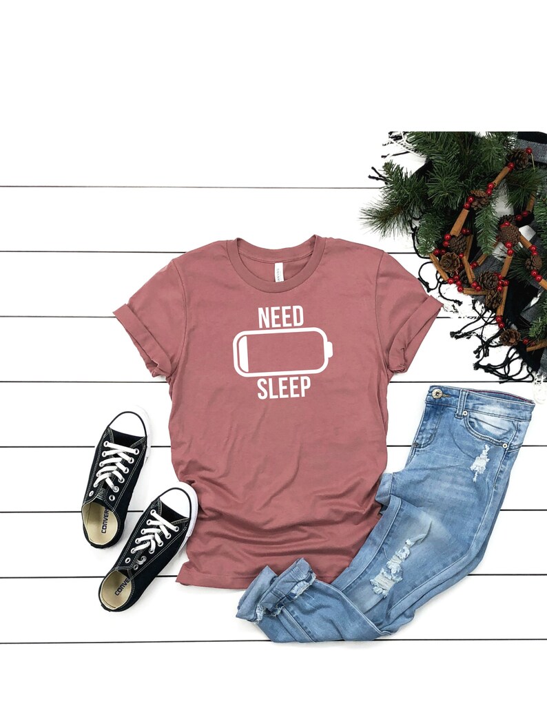 May include: A mauve t-shirt with a white graphic of a battery icon and the text "NEED SLEEP". The shirt is laid out on a white wooden surface with a pair of black sneakers, blue jeans, and a decorative wreath.