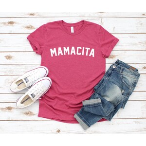 Pu&ograve; includere: Una maglietta rosa con la parola "MAMACITA" stampata in bianco sul davanti. La maglietta &egrave; stesa su una superficie di legno bianco con un paio di scarpe da ginnastica bianche e un paio di jeans blu.
