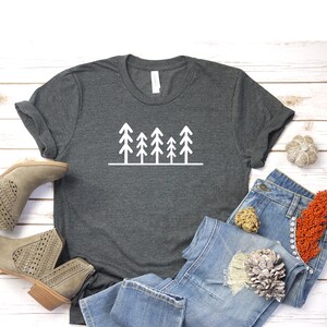 May include: A dark gray t-shirt with a white graphic of a row of five stylized trees. The t-shirt is laid out on a white wooden surface with a pair of brown ankle boots, blue jeans, and a decorative pinecone.