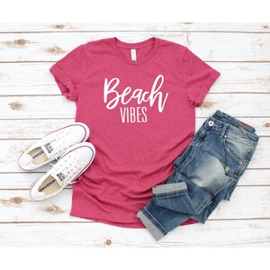 K&ouml;nnte beinhalten: Ein pinkfarbenes T-Shirt mit dem Schriftzug "Beach Vibes" in wei&szlig;en Buchstaben. Das Shirt liegt auf einer wei&szlig;en Holzoberfl&auml;che mit einem Paar wei&szlig;en Turnschuhen und einer blauen Jeans.