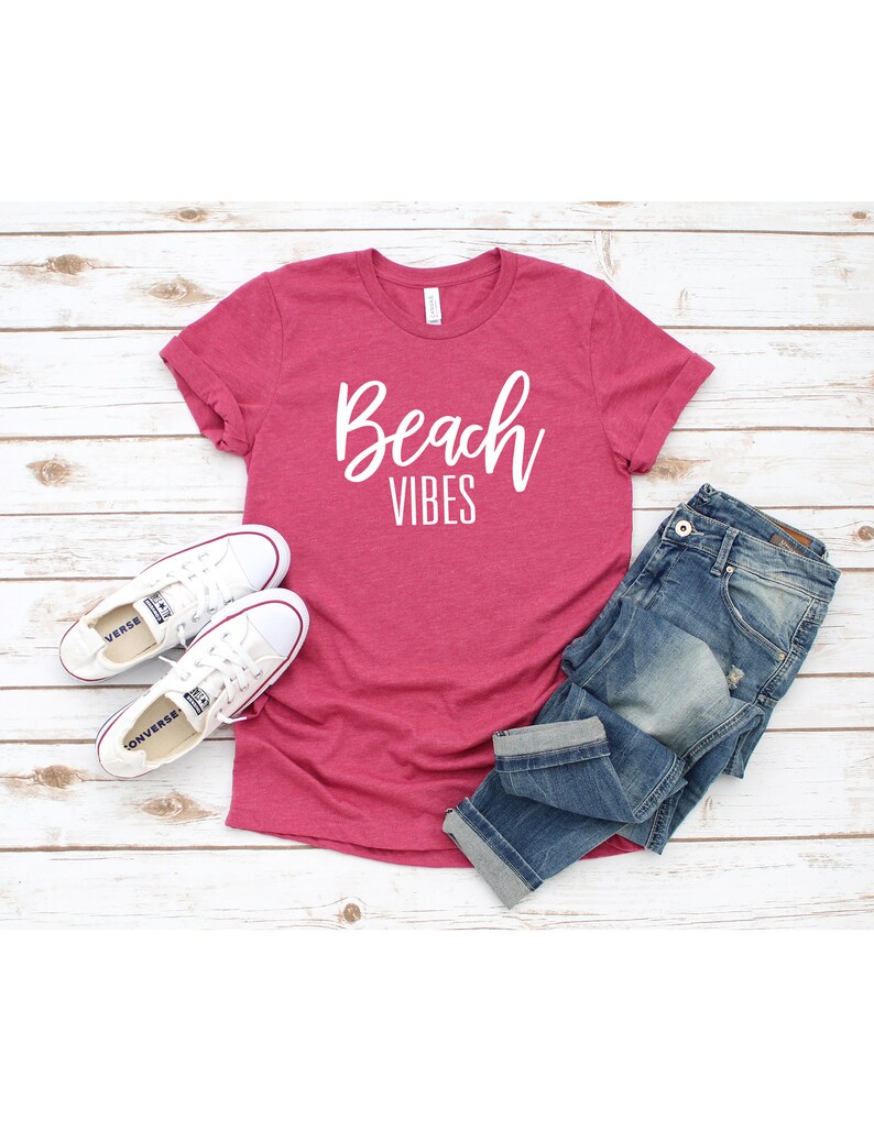 K&ouml;nnte beinhalten: Ein pinkfarbenes T-Shirt mit dem Schriftzug "Beach Vibes" in wei&szlig;en Buchstaben. Das Shirt liegt auf einer wei&szlig;en Holzoberfl&auml;che mit einem Paar wei&szlig;en Turnschuhen und einer blauen Jeans.