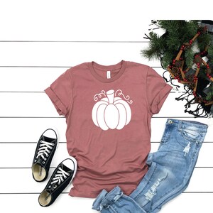 May include: A mauve t-shirt with a white pumpkin graphic. The shirt is laid flat on a white wooden surface with a pair of black converse shoes, blue jeans, and a pine branch with pinecones.