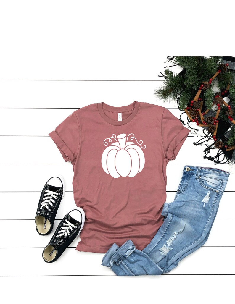 May include: A mauve t-shirt with a white pumpkin graphic. The shirt is laid flat on a white wooden surface with a pair of black converse shoes, blue jeans, and a pine branch with pinecones.