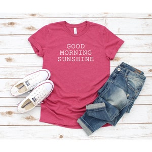 May include: A pink t-shirt with the text "GOOD MORNING SUNSHINE" in white. The shirt is laid out on a white wooden surface with a pair of white sneakers and blue jeans.