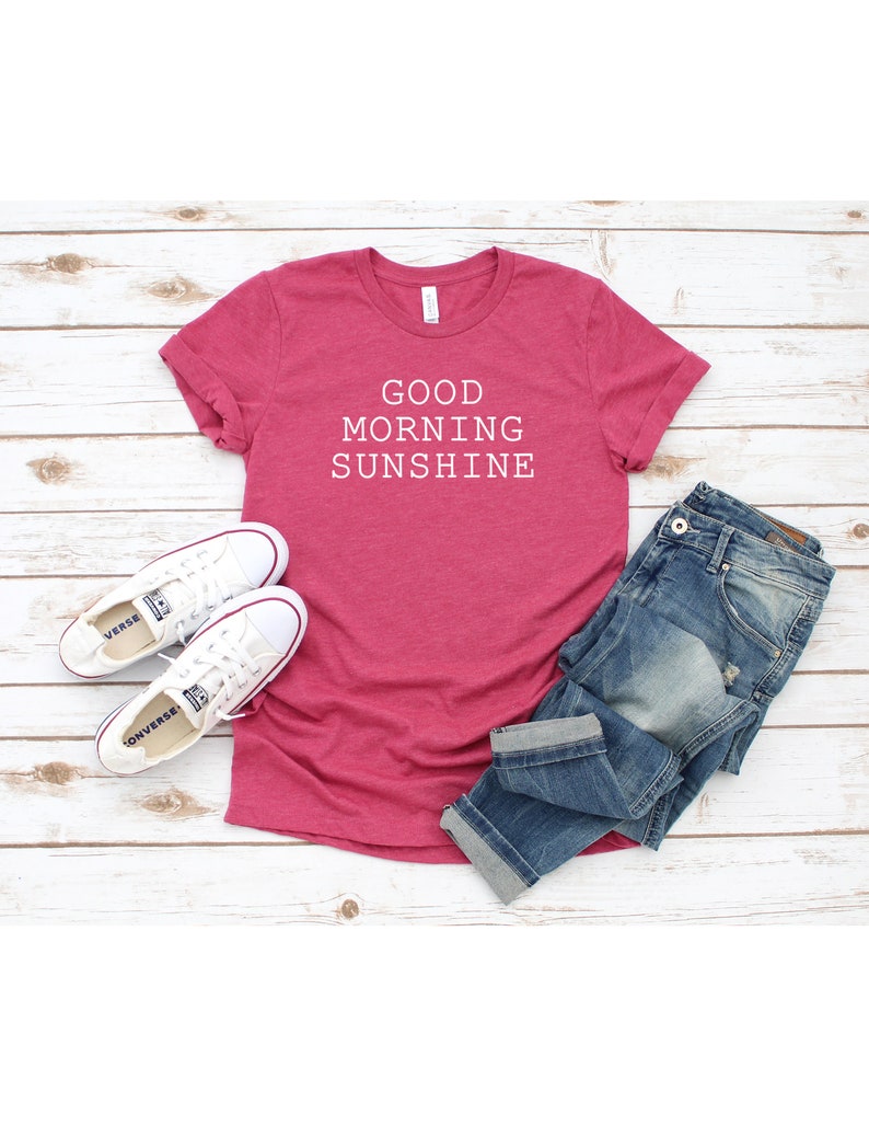 May include: A pink t-shirt with the text "GOOD MORNING SUNSHINE" in white. The shirt is laid out on a white wooden surface with a pair of white sneakers and blue jeans.