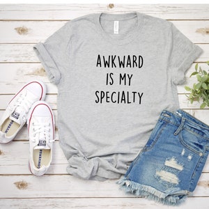 May include: Gray t-shirt with the text "Awkward is my specialty" printed in black. The shirt is laid flat on a white wooden surface with a pair of white sneakers and blue denim shorts.