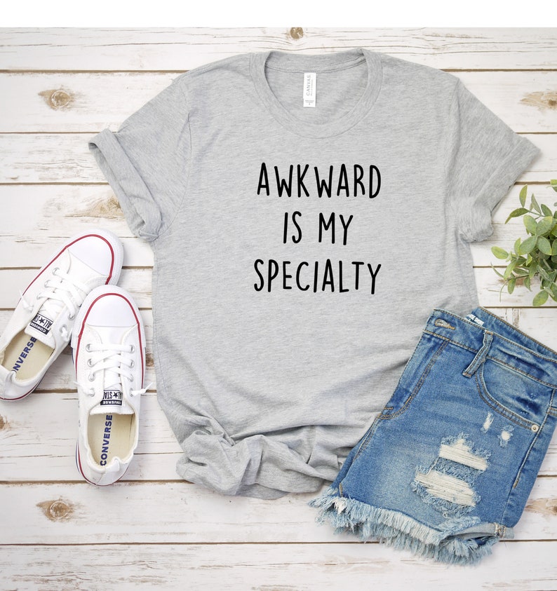 May include: Gray t-shirt with the text "Awkward is my specialty" printed in black. The shirt is laid flat on a white wooden surface with a pair of white sneakers and blue denim shorts.
