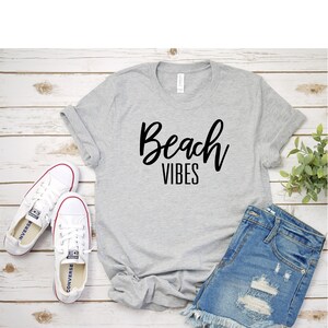 K&ouml;nnte beinhalten: Ein graues T-Shirt mit dem Text "Beach Vibes" in schwarzen Buchstaben. Das Shirt liegt auf einer Holzoberfl&auml;che mit einem Paar wei&szlig;en Sneakers, Jeans-Shorts und einer kleinen Topfpflanze.