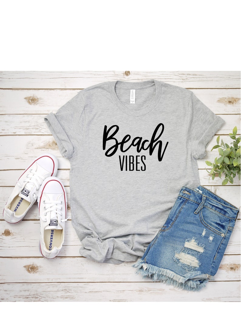 K&ouml;nnte beinhalten: Ein graues T-Shirt mit dem Text "Beach Vibes" in schwarzen Buchstaben. Das Shirt liegt auf einer Holzoberfl&auml;che mit einem Paar wei&szlig;en Sneakers, Jeans-Shorts und einer kleinen Topfpflanze.