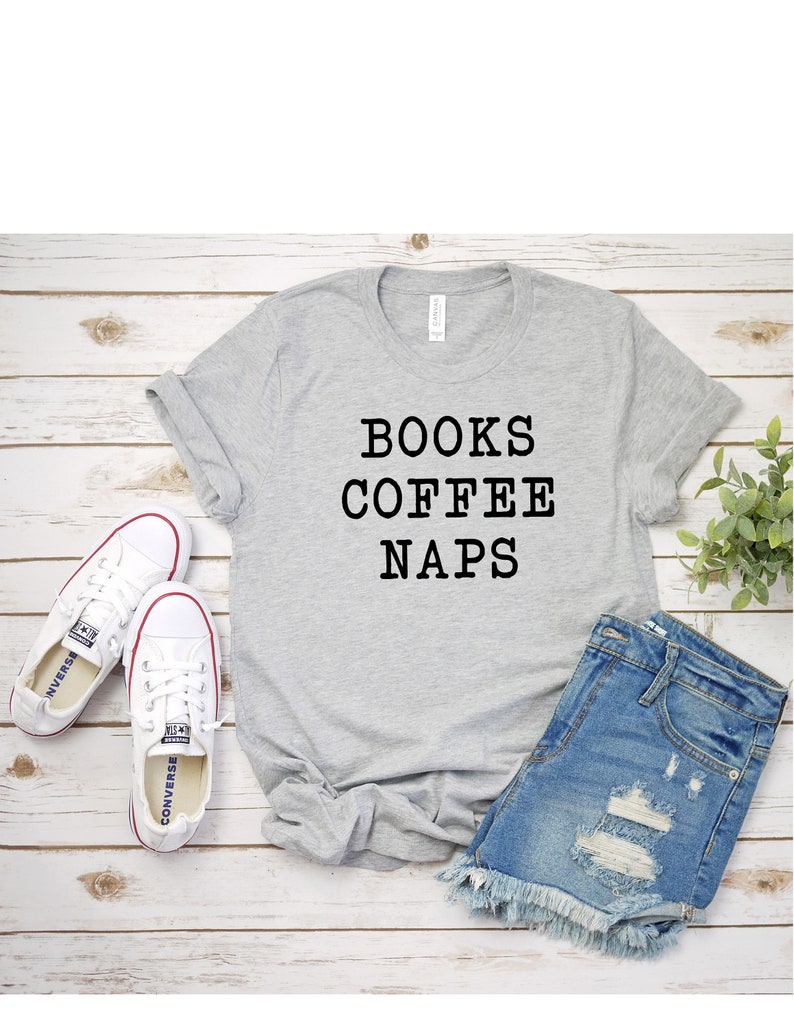 May include: A gray t-shirt with the text "BOOKS COFFEE NAPS" printed in black. The shirt is laid flat on a wooden surface with a pair of white sneakers and denim shorts.
