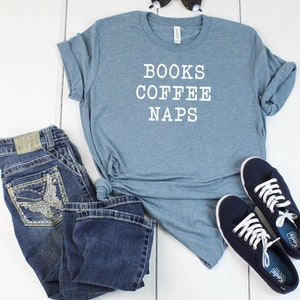 May include: A blue t-shirt with the text "BOOKS COFFEE NAPS" in white. The shirt is paired with blue jeans and navy blue sneakers.