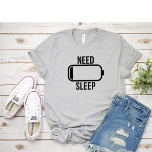 May include: A gray t-shirt with a black graphic of a battery icon and the text "NEED SLEEP". The shirt is laid flat on a wooden surface with a pair of white sneakers and denim shorts.