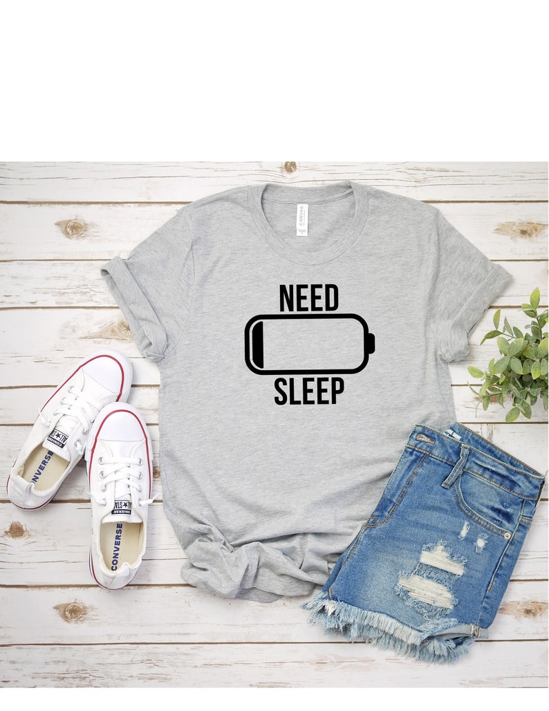 May include: A gray t-shirt with a black graphic of a battery icon and the text "NEED SLEEP". The shirt is laid flat on a wooden surface with a pair of white sneakers and denim shorts.