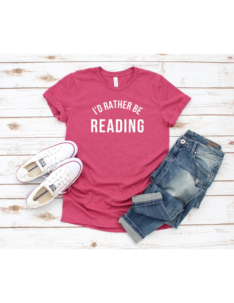 May include: A pink t-shirt with the text "I'd rather be reading" in white. The shirt is on a white wooden background with a pair of white sneakers and blue jeans.
