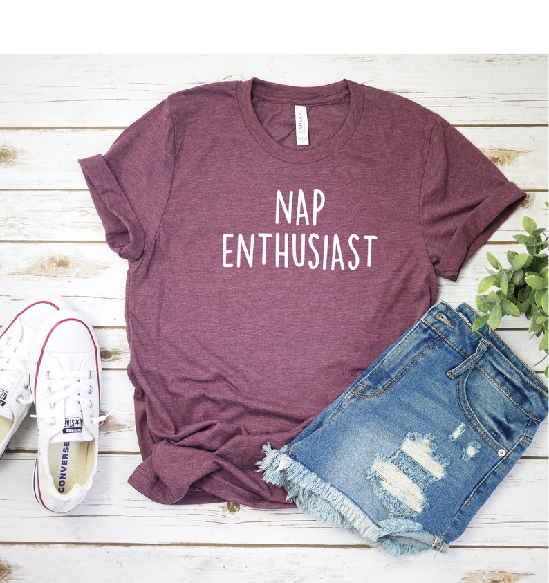 May include: A maroon t-shirt with the text "NAP ENTHUSIAST" in white lettering. The shirt is laying on a white wooden surface with a pair of white sneakers and a pair of blue denim shorts.
