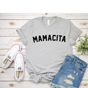 Pu&ograve; includere: Una maglietta grigia con la scritta "MAMACITA" stampata in nero. La maglietta &egrave; appoggiata su una superficie di legno bianco con un paio di scarpe da ginnastica bianche, un paio di pantaloncini di jeans e una piccola pianta verde.