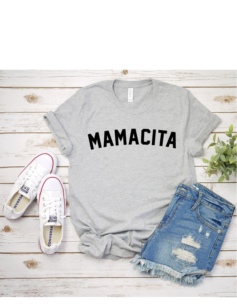 Pu&ograve; includere: Una maglietta grigia con la scritta "MAMACITA" stampata in nero. La maglietta &egrave; appoggiata su una superficie di legno bianco con un paio di scarpe da ginnastica bianche, un paio di pantaloncini di jeans e una piccola pianta verde.