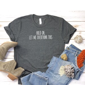 May include: A dark gray t-shirt with the text "Hold on, let me overthink this" printed in white. The shirt is laid out on a white wooden surface with a pair of light brown ankle boots, blue jeans, and decorative items.