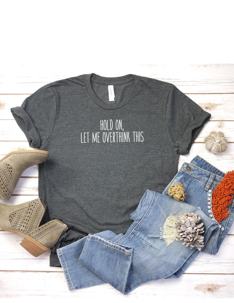 May include: A dark gray t-shirt with the text "Hold on, let me overthink this" printed in white. The shirt is laid out on a white wooden surface with a pair of light brown ankle boots, blue jeans, and decorative items.