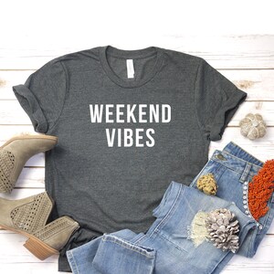 May include: A dark gray t-shirt with the text "Weekend Vibes" in white lettering. The shirt is laid out on a white wooden surface with a pair of light brown ankle boots, blue jeans, and decorative items.