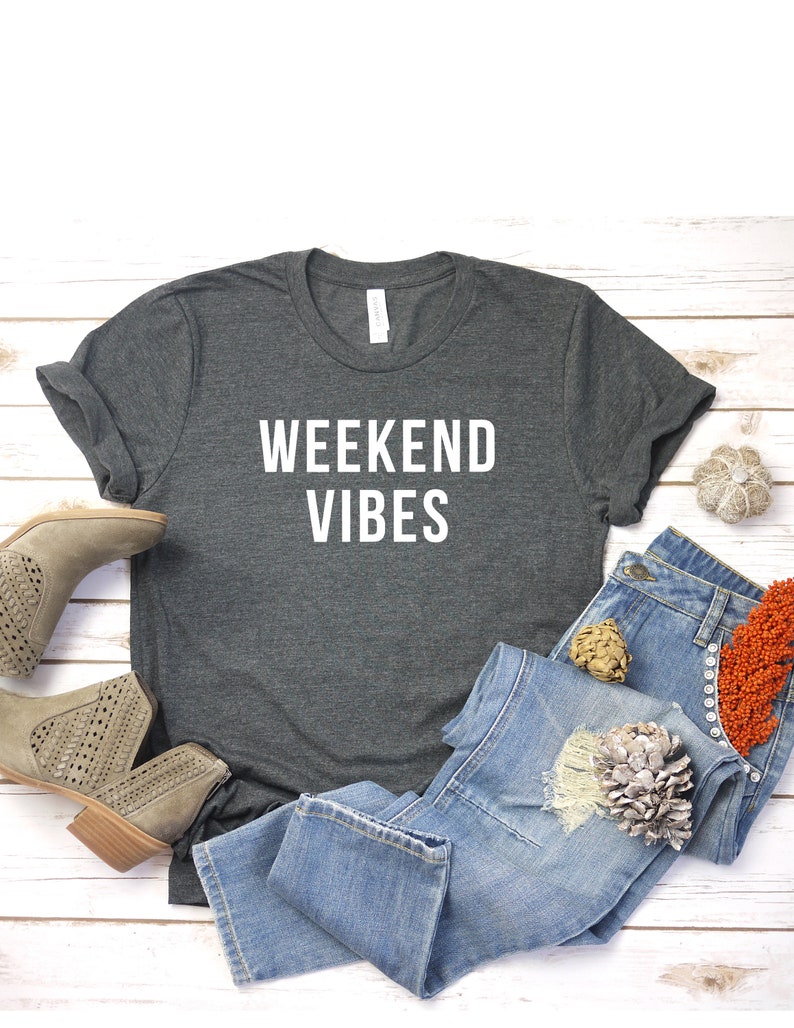 May include: A dark gray t-shirt with the text "Weekend Vibes" in white lettering. The shirt is laid out on a white wooden surface with a pair of light brown ankle boots, blue jeans, and decorative items.