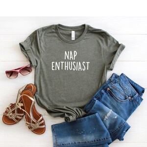 May include: A green t-shirt with the text "NAP ENTHUSIAST" in white lettering. The shirt is laid out on a white wooden surface with a pair of brown sandals and blue jeans.