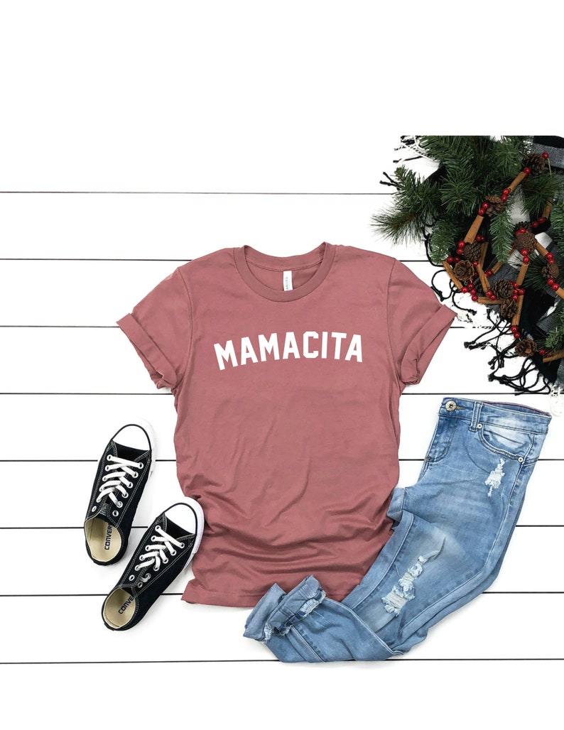 Pu&ograve; includere: Una t-shirt color malva con la scritta "MAMACITA" stampata in bianco sul davanti. La t-shirt &egrave; abbinata a jeans blu chiaro strappati e sneakers di tela nere.