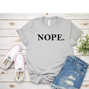 May include: A gray t-shirt with the word "NOPE." printed in black on the front. The shirt is paired with denim shorts and white sneakers.