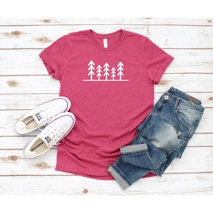 May include: A pink t-shirt with a white graphic of a row of stylized pine trees. The shirt is laying on a white wooden surface with a pair of white sneakers and blue jeans.