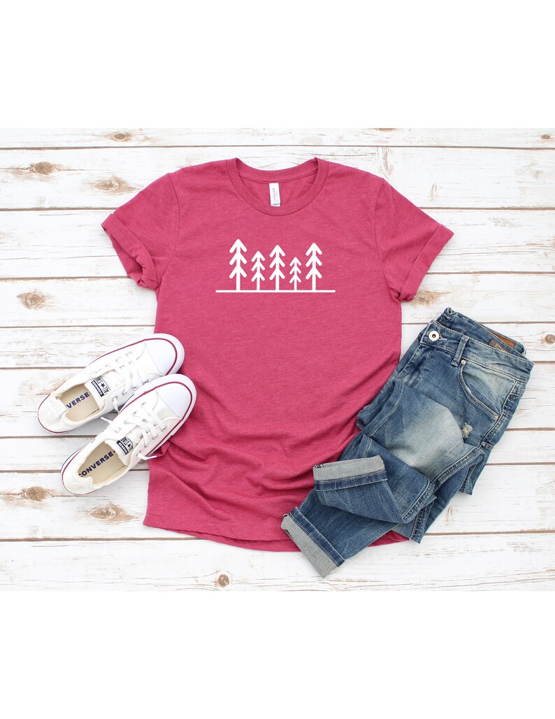 May include: A pink t-shirt with a white graphic of a row of stylized pine trees. The shirt is laying on a white wooden surface with a pair of white sneakers and blue jeans.