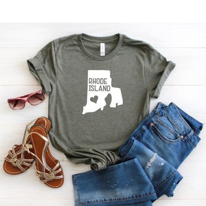 K&ouml;nnte beinhalten: Ein gr&uuml;nes T-Shirt mit einer wei&szlig;en Umrandung des Bundesstaates Rhode Island und dem Text "Rhode Island" mit einem wei&szlig;en Herzen innerhalb der Bundesstaatenumrandung.