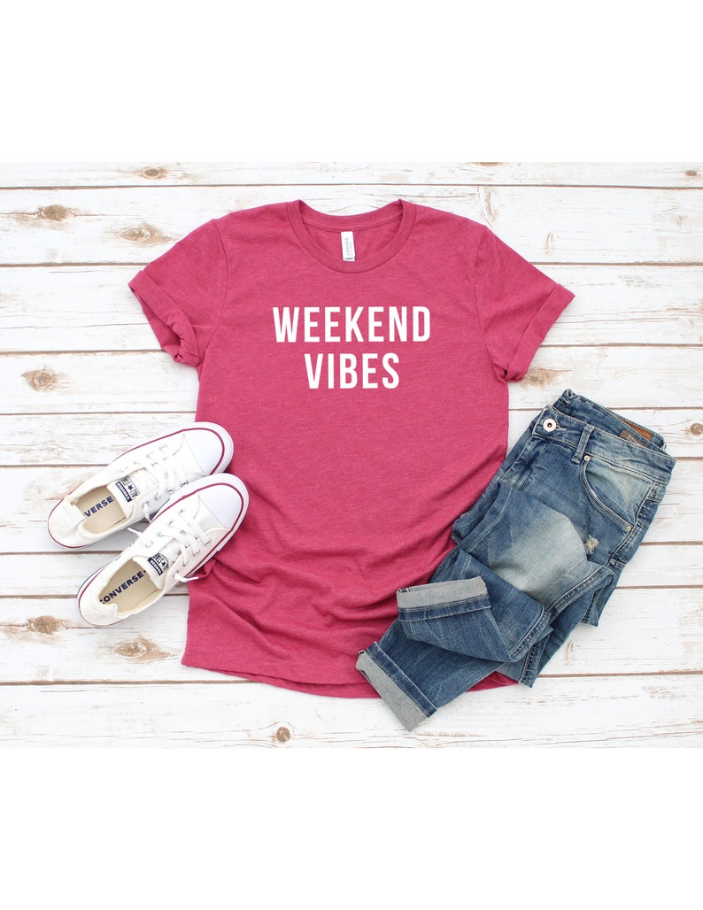 May include: A pink t-shirt with the text "Weekend Vibes" in white lettering. The shirt is laid flat on a white wooden surface with a pair of white sneakers and blue jeans.