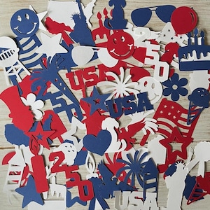 May include: A collection of red, white, and blue paper cutouts. The cutouts include stars, the Statue of Liberty, the word "USA", smiley faces, an American flag, and a birthday cake. The shapes are scattered across a light wood surface.