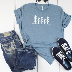 May include: A blue t-shirt with a white graphic of a row of trees. The t-shirt is paired with blue jeans and blue sneakers.
