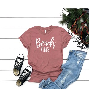 K&ouml;nnte beinhalten: Ein mauvefarbenes T-Shirt mit dem Schriftzug "Beach Vibes" in wei&szlig;er Schrift. Das T-Shirt liegt auf einer wei&szlig;en Holzoberfl&auml;che mit einem Paar schwarzer Turnschuhe, einer blauen Jeans und einem gr&uuml;nen Kranz mit braunen Tannenzapfen.