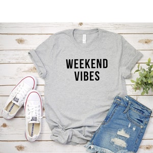 May include: A gray t-shirt with the text "Weekend Vibes" printed in black. The t-shirt is laid out on a wooden surface with a pair of white sneakers and denim shorts.