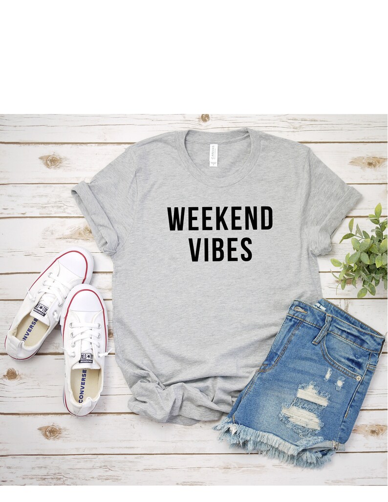 May include: A gray t-shirt with the text "Weekend Vibes" printed in black. The t-shirt is laid out on a wooden surface with a pair of white sneakers and denim shorts.