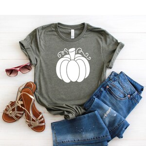 May include: A green t-shirt with a white pumpkin graphic. The pumpkin has a stem and curly vines. The t-shirt is laid out on a white wooden surface with a pair of brown sandals and blue jeans.