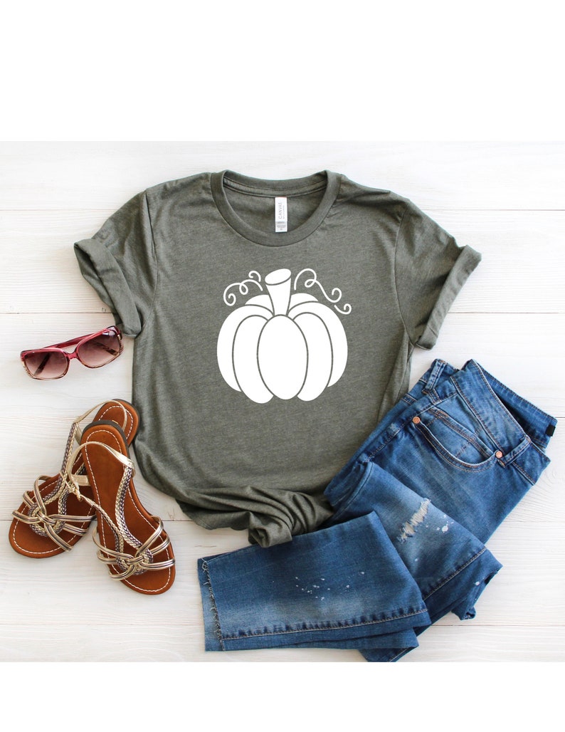 May include: A green t-shirt with a white pumpkin graphic. The pumpkin has a stem and curly vines. The t-shirt is laid out on a white wooden surface with a pair of brown sandals and blue jeans.