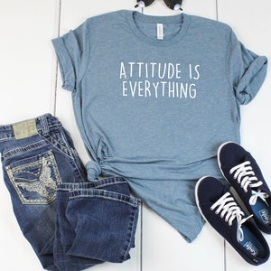 May include: A blue t-shirt with the text "Attitude is Everything" in white lettering. The t-shirt is paired with blue jeans and navy blue sneakers.