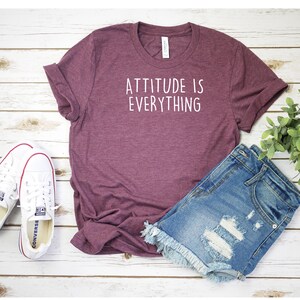 May include: A maroon t-shirt with the text "Attitude is Everything" in white lettering. The t-shirt is laying on a white wooden surface with a pair of white sneakers and denim shorts.