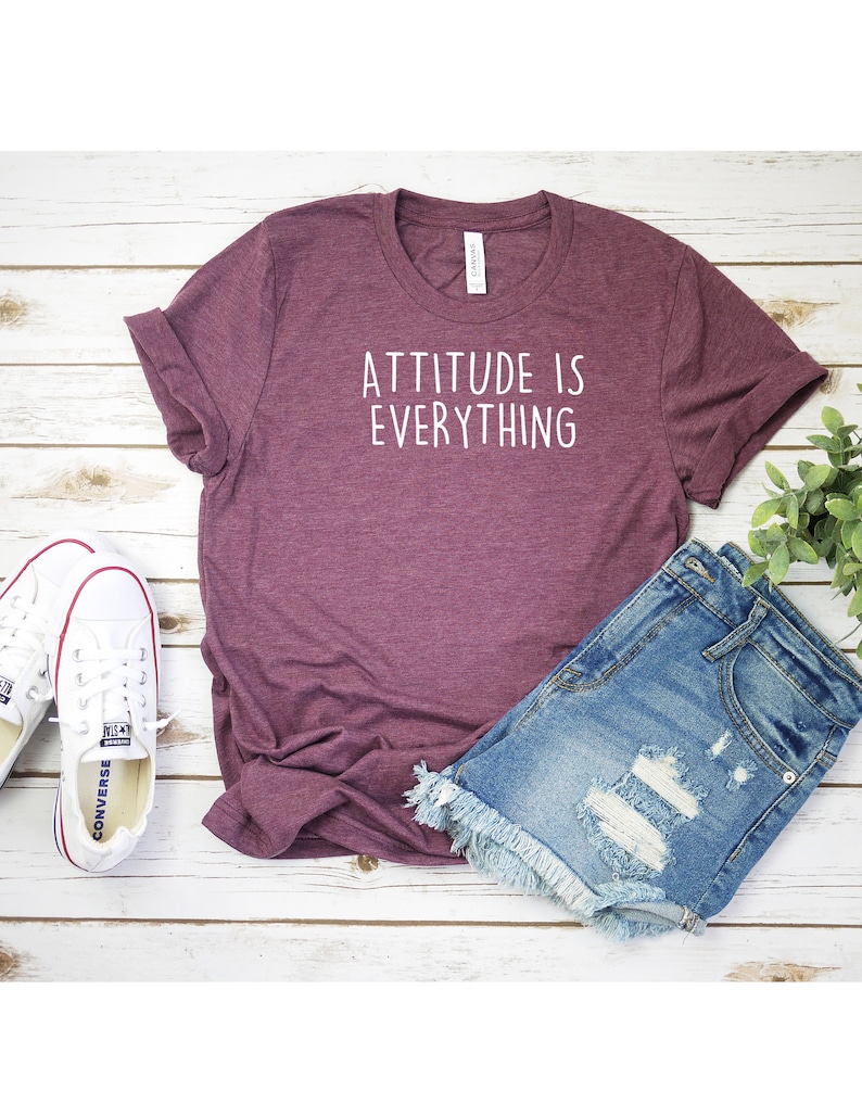 May include: A maroon t-shirt with the text "Attitude is Everything" in white lettering. The t-shirt is laying on a white wooden surface with a pair of white sneakers and denim shorts.