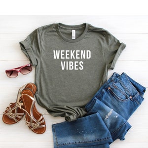 May include: A green t-shirt with the text "Weekend Vibes" in white lettering. The shirt is laid out on a white wooden surface with a pair of brown sandals and blue jeans.
