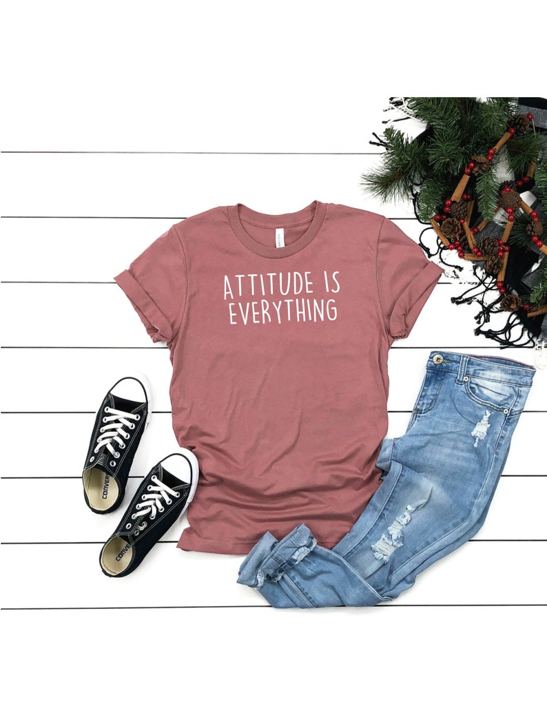 May include: A mauve t-shirt with the text "ATTITUDE IS EVERYTHING" in white lettering. The t-shirt is laid flat on a white wooden surface with a pair of black Converse sneakers and blue jeans with distressed details.