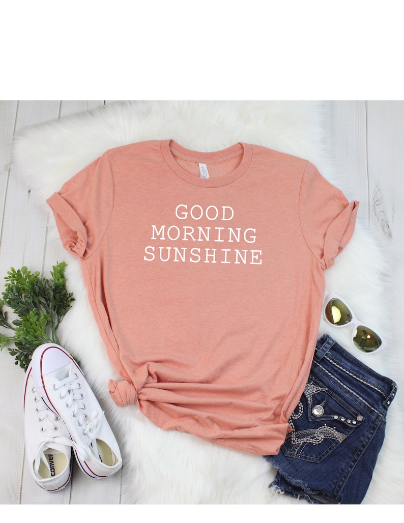 May include: A peach-colored t-shirt with the text "GOOD MORNING SUNSHINE" printed in white. The shirt is laid out on a white fuzzy surface with a pair of white sneakers, a pair of blue denim shorts, and a pair of sunglasses.