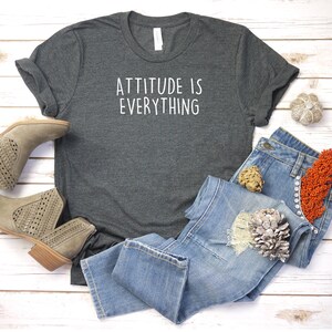 May include: A dark gray t-shirt with the text "ATTITUDE IS EVERYTHING" printed in white on the front. The shirt is laid flat on a white wooden surface with a pair of brown ankle boots and blue jeans.