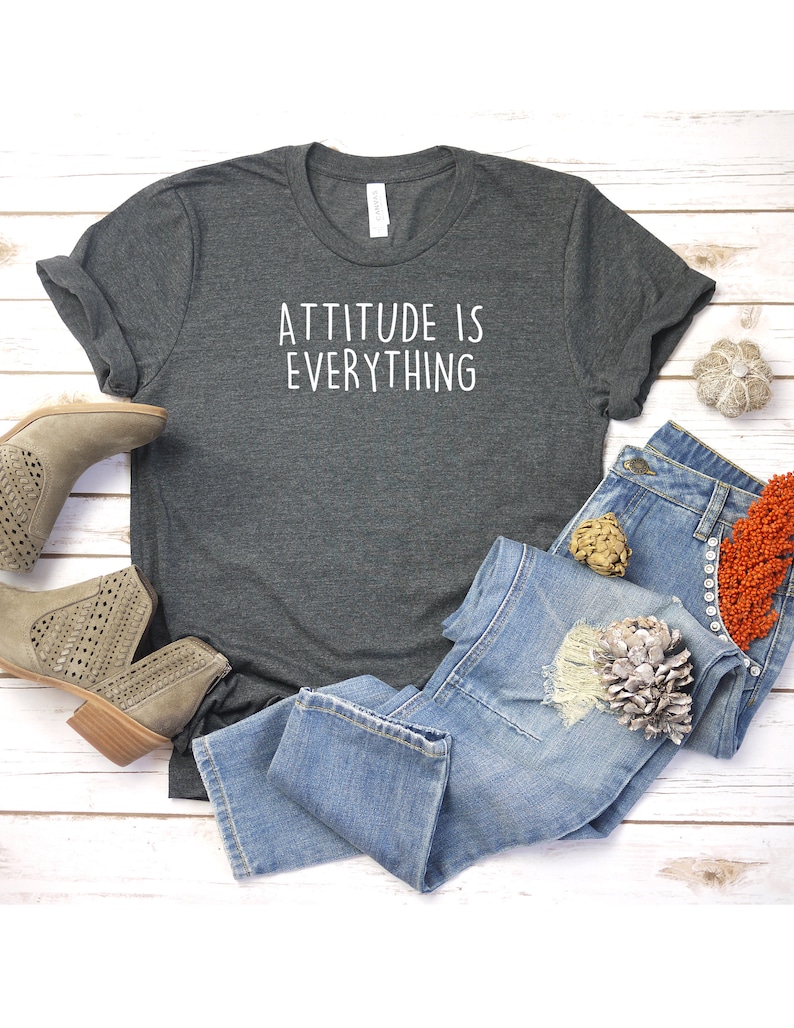 May include: A dark gray t-shirt with the text "ATTITUDE IS EVERYTHING" printed in white on the front. The shirt is laid flat on a white wooden surface with a pair of brown ankle boots and blue jeans.