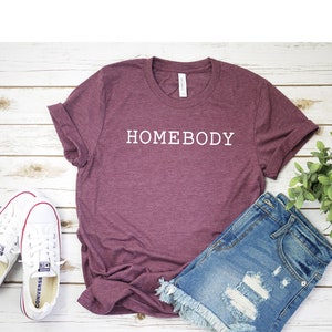 Pode incluir: Uma camiseta vinho com o texto "HOMEBODY" em letras brancas. A camiseta est&aacute; colocada sobre uma superf&iacute;cie de madeira branca com um par de t&ecirc;nis brancos e shorts jeans.