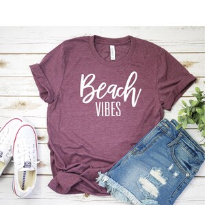 K&ouml;nnte beinhalten: Ein maroonfarbenes T-Shirt mit dem Text "Beach Vibes" in wei&szlig;en Buchstaben. Das Shirt liegt flach auf einer wei&szlig;en Holzoberfl&auml;che mit einem Paar wei&szlig;en Turnschuhen und einer Jeans-Shorts.